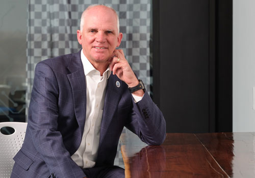 Dr. Paul Illich is in a navy blue suit jacket and white dress shirt, sitting at a table he built for the Sandhills Global Technology Center. The background shows a contemporary office interior with glass and neutral tones.