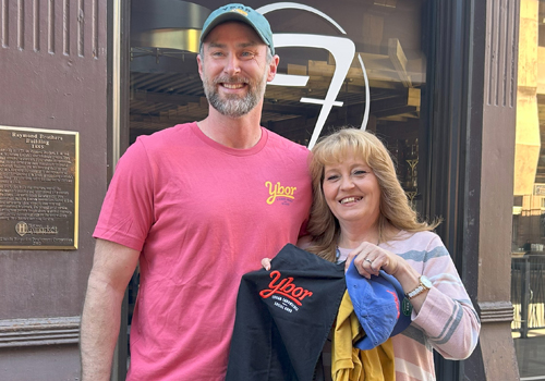 Bill Radtke (left) and Becky Olson smile outside Fleetwood as Radtke gives Olson Ybor branded merchandise including a black apron, yellow t-shirt and blue hat.