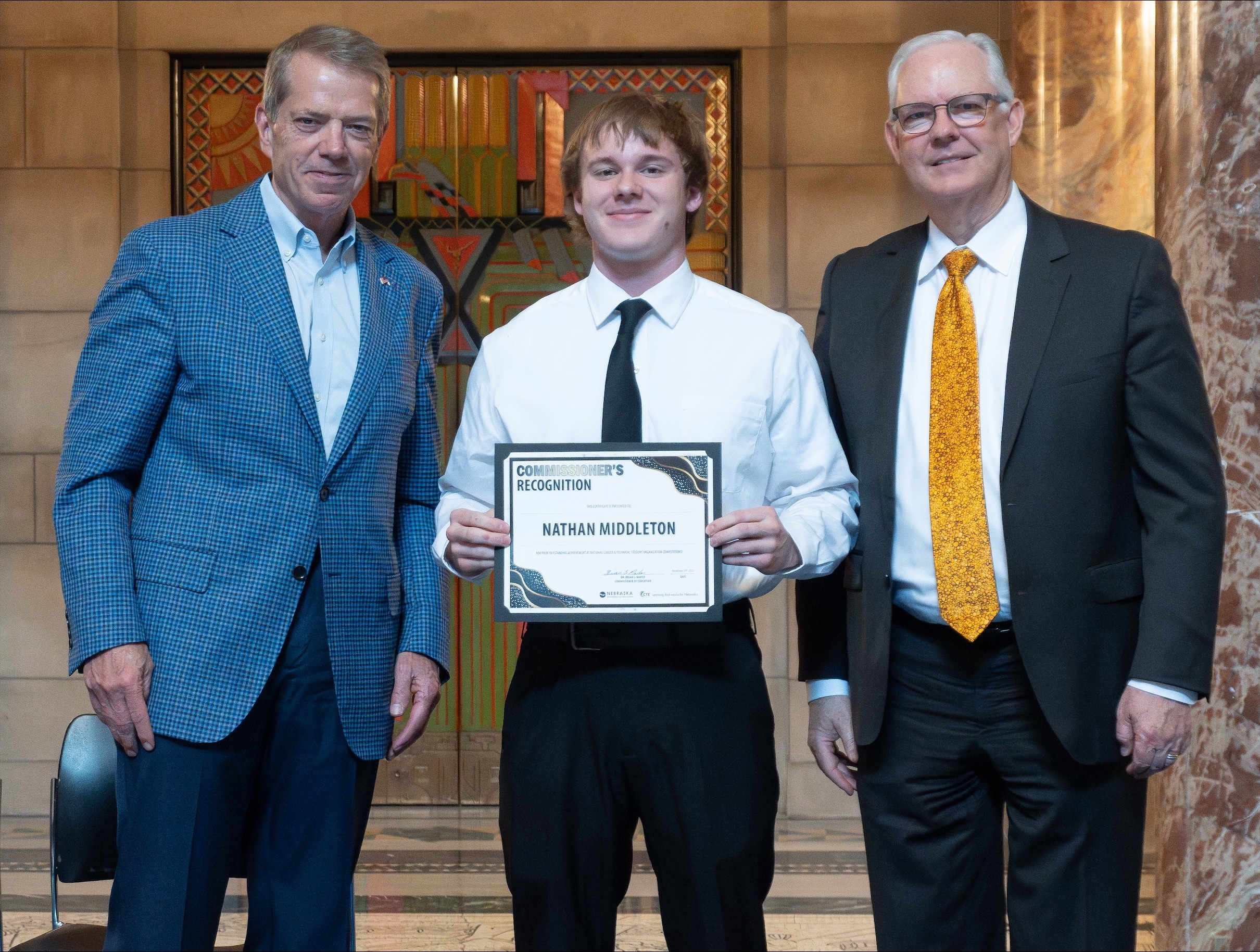 Jim Pillen and Dr. Brian Maher stand together posing for a photo in the capitol. In the center, Nathan Middleton, holds a framed 
