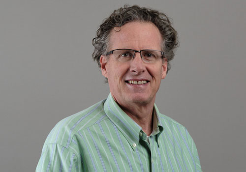 Phip Ross is smiling wearing a light green striped button-up shirt against a neutral gray background. 