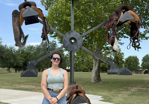 Emma Skala stands in front of a large metal welded wheel she designed to hold saddles