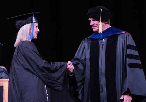 Crissy Murphy (left) in a black cap and gown shakes hands with  SCC President Dr. Paul Illich wearing formal academic attire with blue trim during the May 14, 2025 graduation ceremony on a darkened stage.