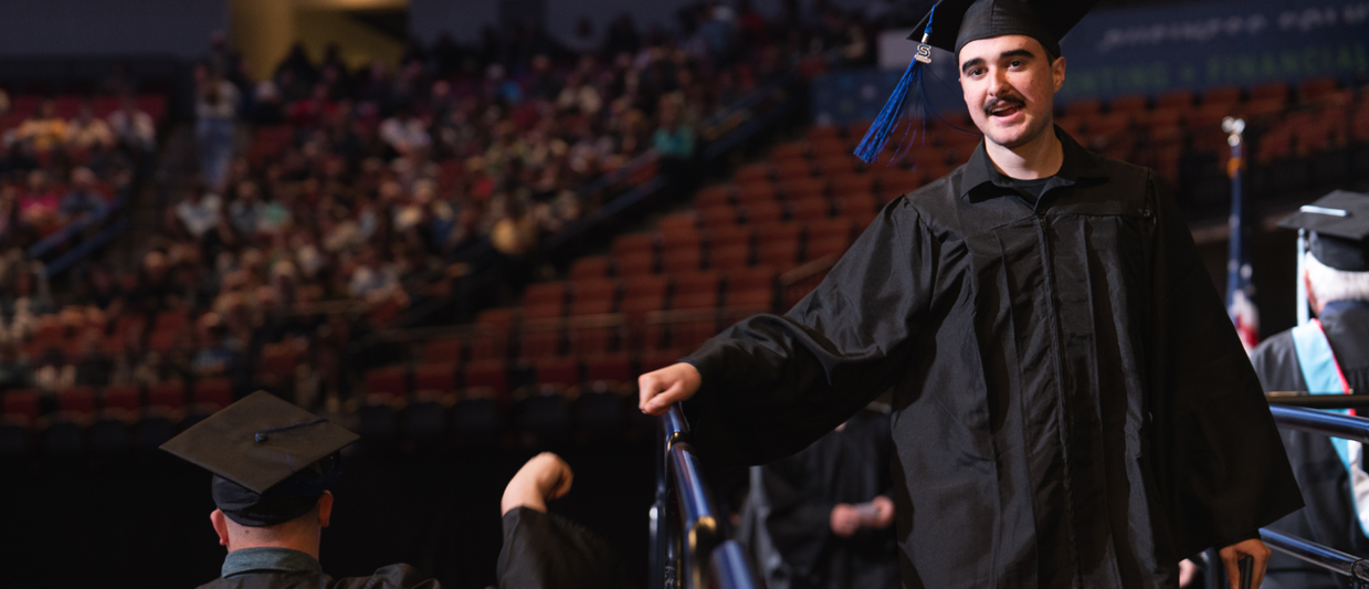 A male graduate in a black cap and gown smiles while walking off the stage at the SCC’s spring graduation, fist-bumping another graduate as they pass.