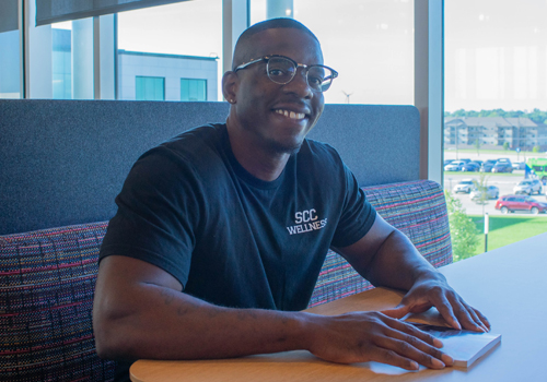 Ricky Sanders is wearing glasses and a black “SCC Wellness” t-shirt, smiling while seated at a table in the library with large windows showing the campus in the background. 
