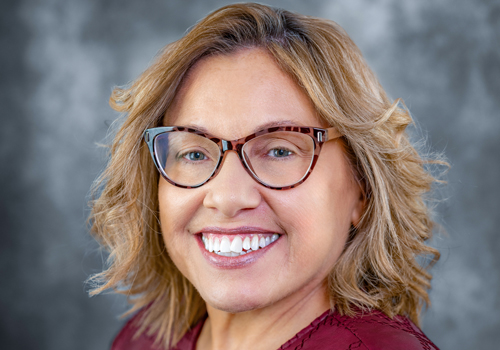 Dawn-Renee Smith smiling in a maroon top with tortoiseshell cat-eye glasses in front of a gray background.  
