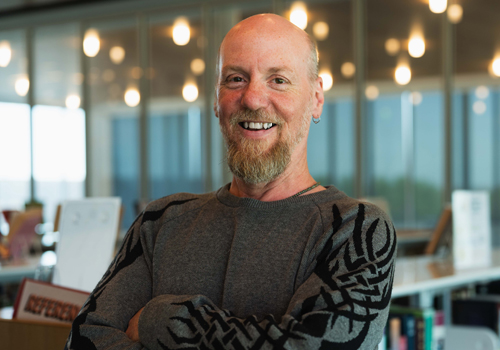 Chad Finch smiles with arms crossed wearing a gray sweater with a black tribal-style pattern and a reddish-blond beard in the library. 