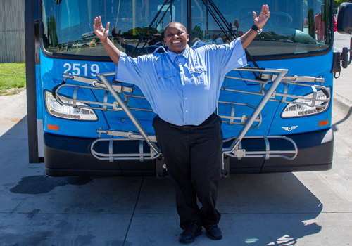 Jessica Whittaker is in a blue Omaha Regional Transit Authority uniform and stands with her arms raised joyfully in front of a bus, smiling widely on a sunny day.