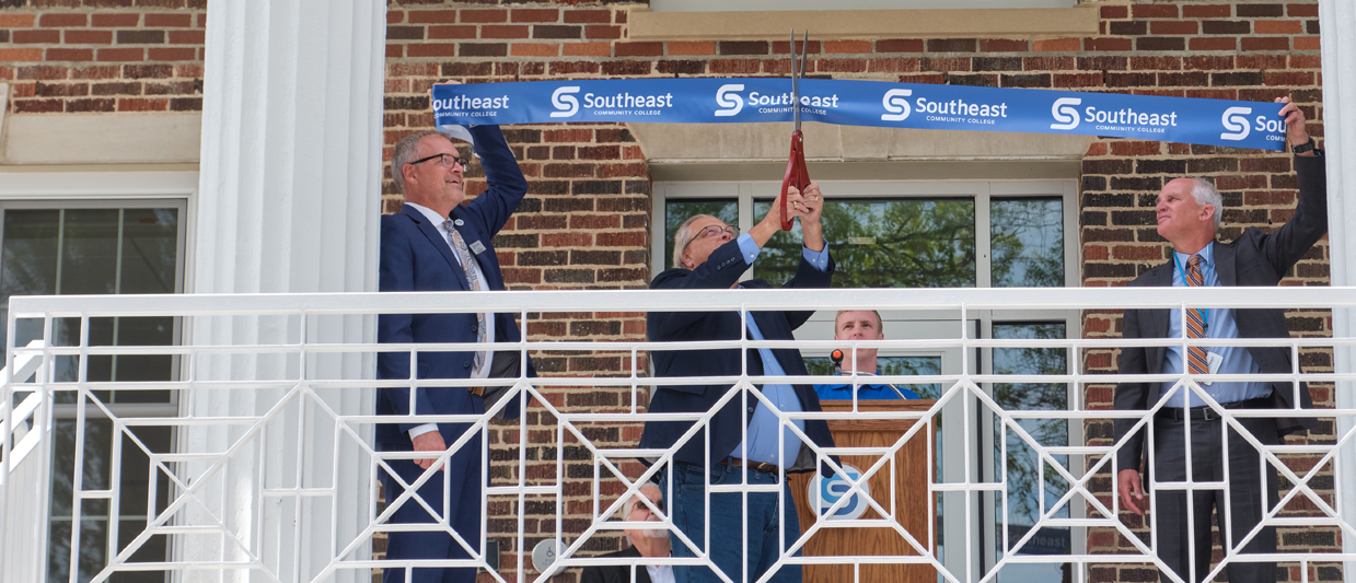 Jon Kisby, Ed Koster and Dr. Paul Illich cut the blue SCC ribbon at Nebraska Hall. All three men are in blue suits. 