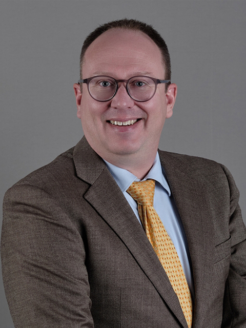 Professional headshot of Matthew Haden wearing round glasses, a brown suit jacket, light blue dress shirt, and yellow patterned tie, smiling at the camera against a gray background.