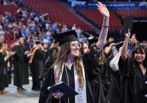 A female graduate in cap and gown waves at her family. 